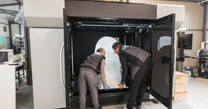 Two workers in black jackets and pants lifting out a large, egg-shaped print from a large-format 3D printer.