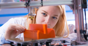 A woman in a white laboratory coat checking the progress of her orange 3D-printed item in her 3D printer.