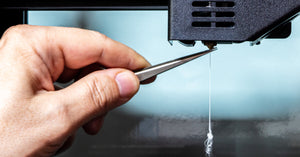 A hand using a pair of tweezers to pull a clogged strand of filament out of the nozzle of a 3D printer.