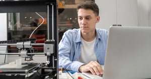 A young man using his laptop to control a 3D printer. The 3D printer is in the middle of the printing process.