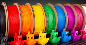 A row of 3D printer filaments arranged in rainbow order—black, red, orange, yellow, green, blue, purple, pink, and white.