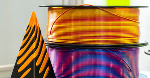 A black and orange 3D-printed object resting on a table next to two rolls of differently colored 3D printer filaments.