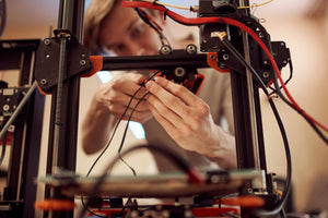 A young person inspecting and working hard on the wiring inside a black and red 3D printer. The person holds a tool.