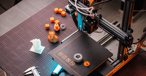 A black and orange 3D printer sits on a workbench. There are orange, gray, and white items near the printer.